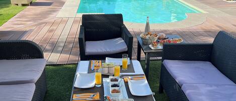 A poolside dining area with wicker furniture, a table set for breakfast, and a view of a swimming pool and hills in the background.