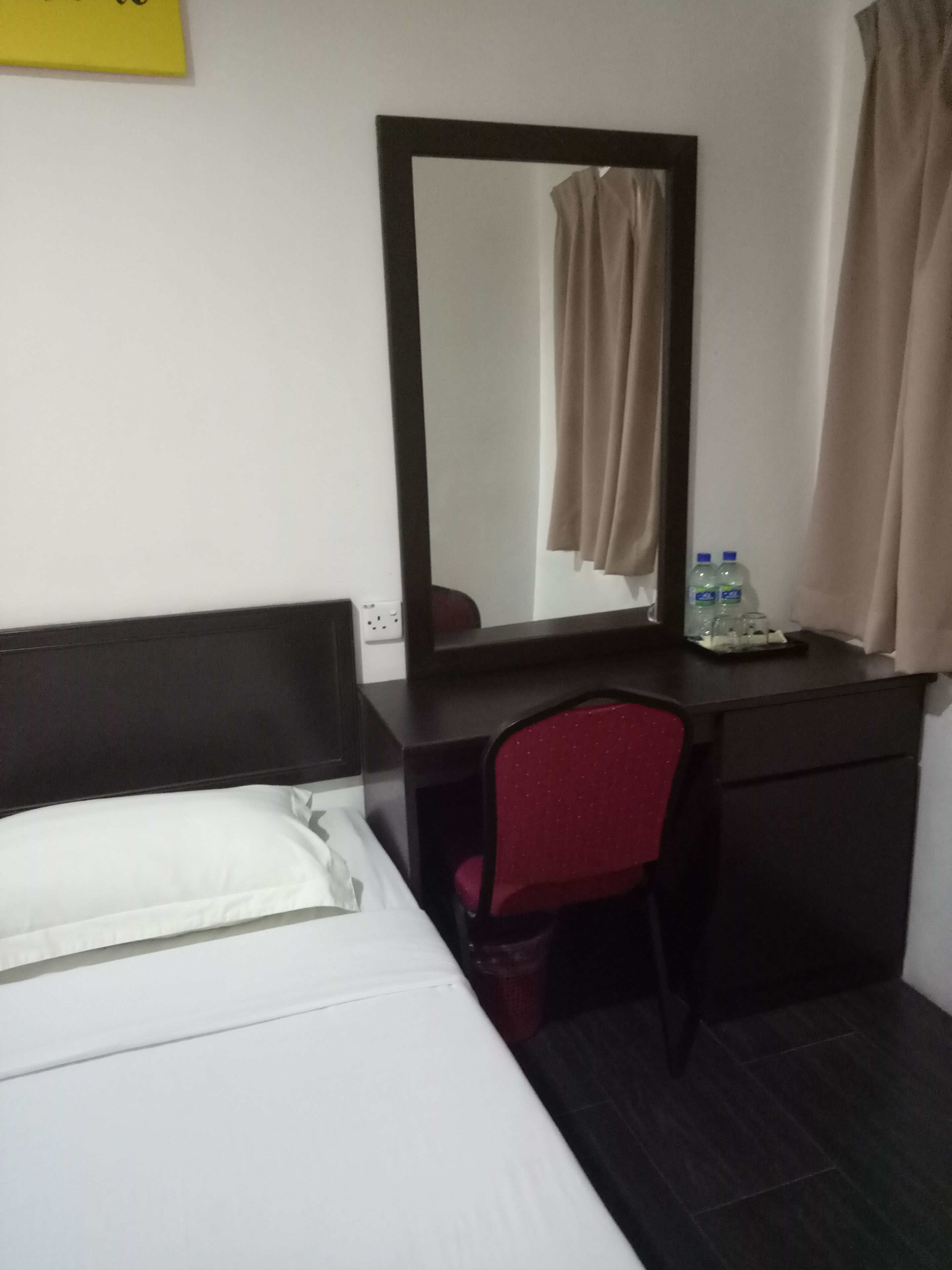 A hotel room with a bed, a desk with a chair, a mirror, a water bottle, and a curtain.