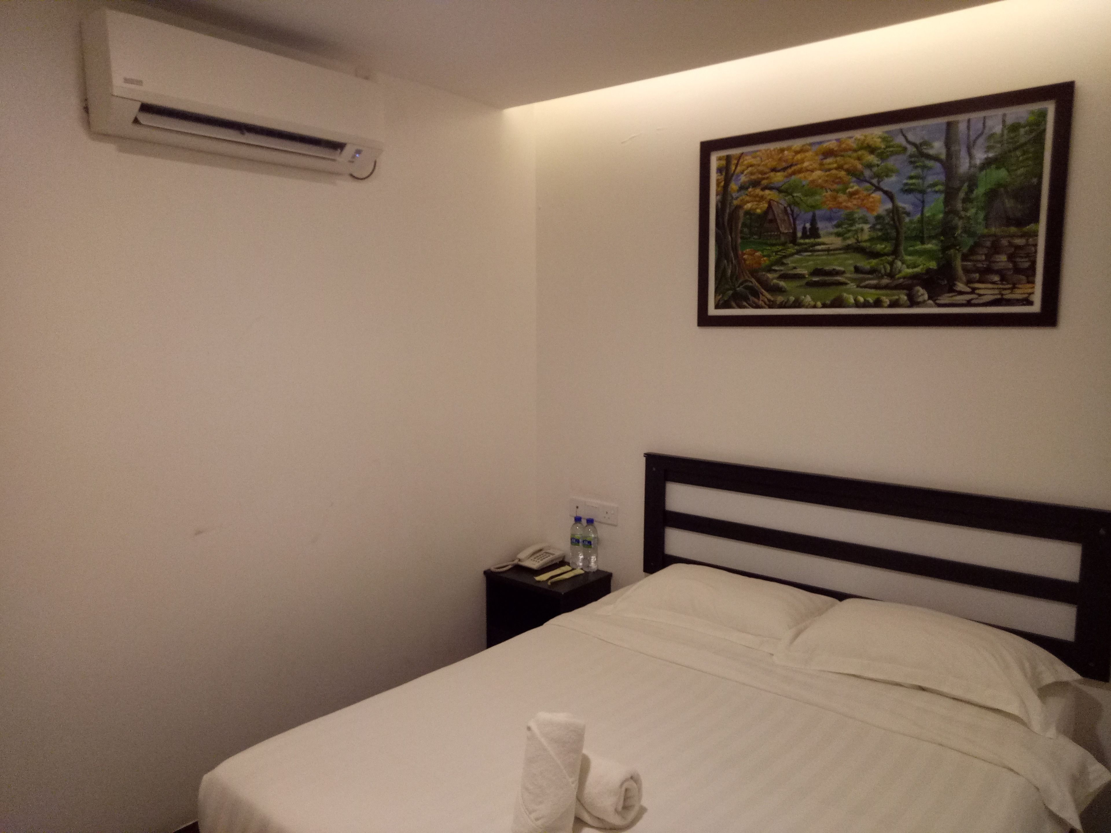 Una cama bien hecha con sábanas blancas, un cuadro de un paisaje boscoso en la pared, y una mesita de noche con un teléfono y botellas de agua.