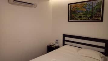 Una cama bien hecha con sábanas blancas, un cuadro de un paisaje boscoso en la pared, y una mesita de noche con un teléfono y botellas de agua.