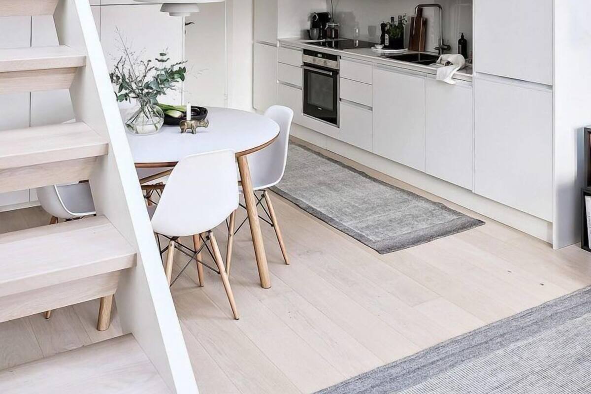Une cuisine moderne avec des meubles blancs, un coin repas doté d’une table et de chaises blanches, et un escalier menant à une chambre située en mezzanine.