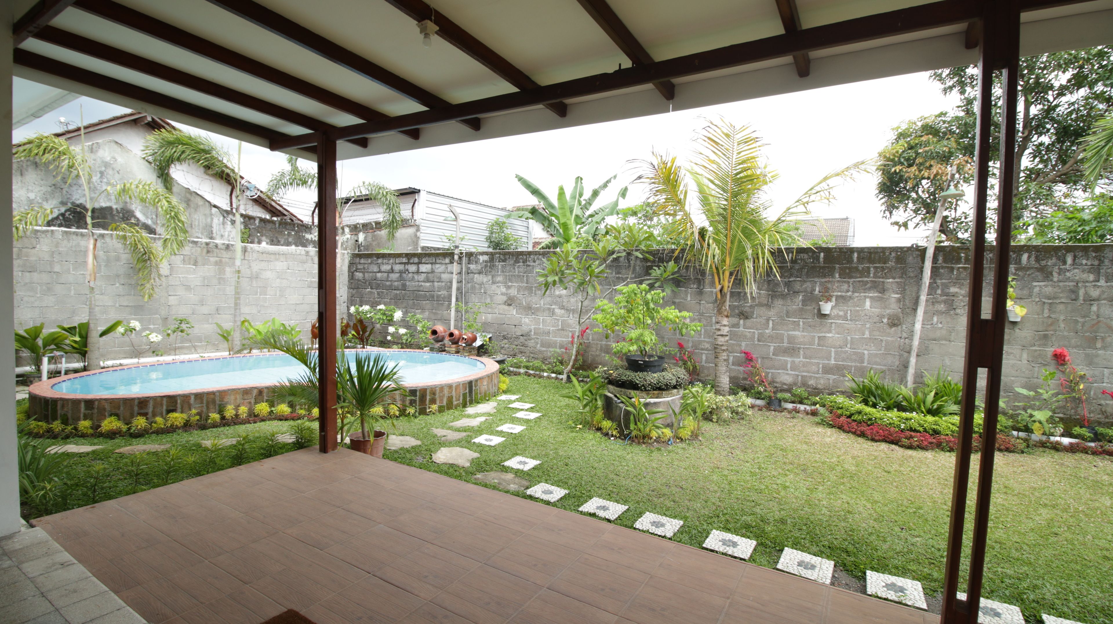 A covered patio with a swimming pool, surrounded by a garden with various plants and a stone wall.