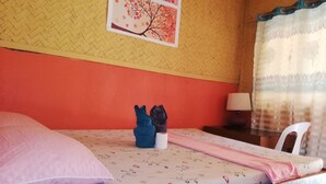 A bed with a pink pillow, a blue and black cushion, and a pink blanket. There is a wall with a woven pattern and a framed picture of a tree. A lamp and a chair are visible in the corner. - Forest Inn (El Nido)
