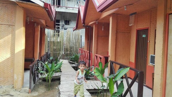 A narrow corridor with a wooden walkway, surrounded by buildings with red railings and a bamboo fence.