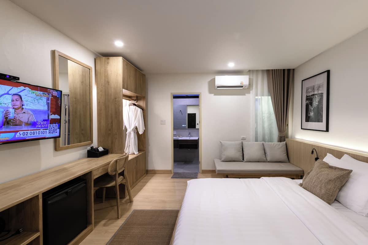 A hotel room with a bed, a desk with a TV, a wardrobe, a sofa, and an air conditioning unit.