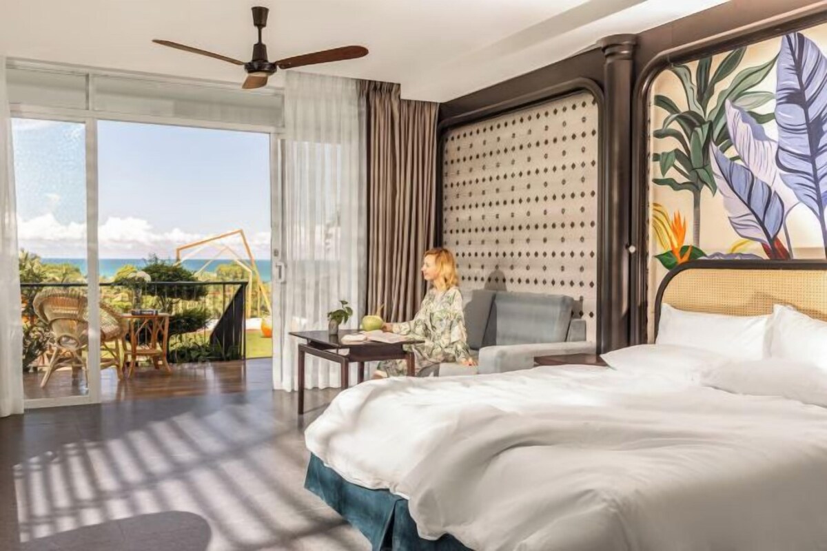 Habitación de hotel con una cama grande, un balcón con vista al océano, un ventilador de techo y una mesa con una planta.