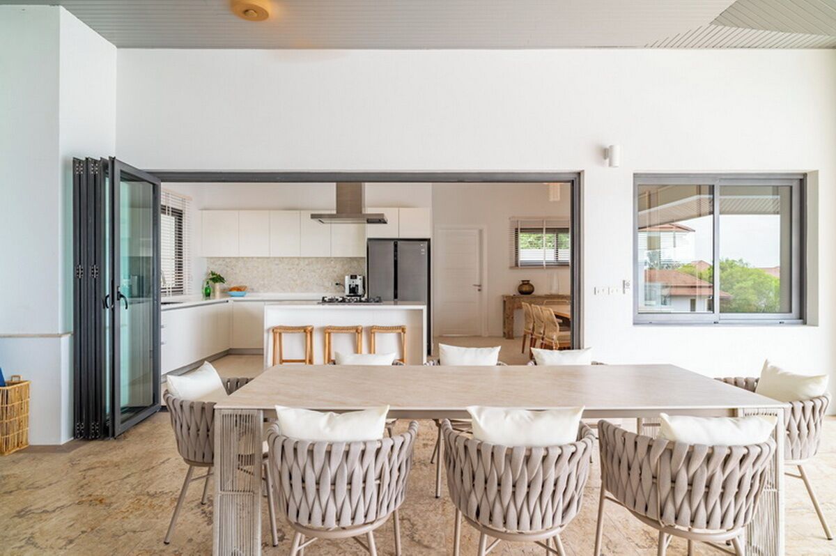 A modern kitchen with white cabinets, a central island, and a dining area with a long table and wicker chairs.