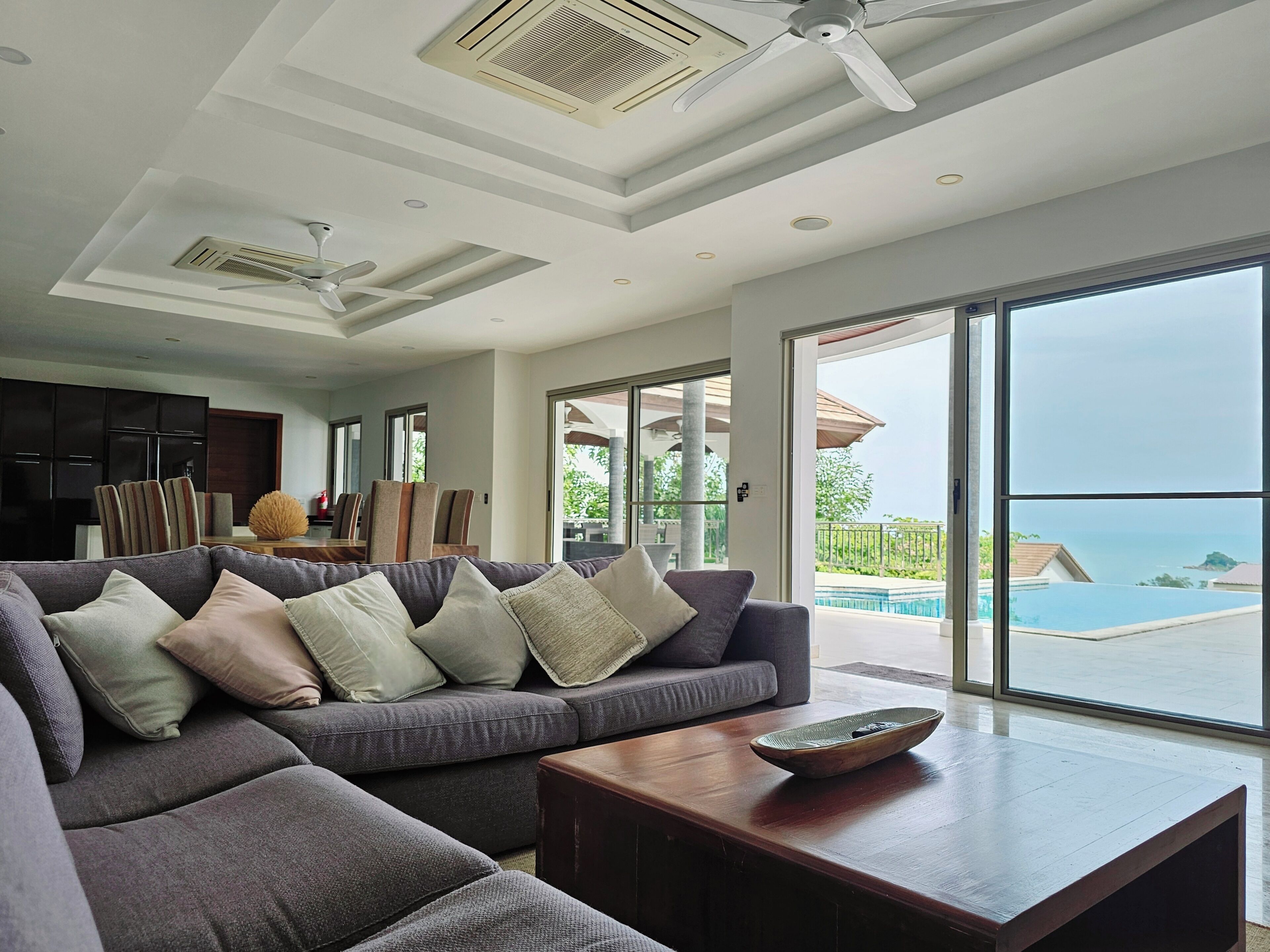 A modern living room with a grey sectional sofa, a wooden coffee table, and a large sliding glass door leading to a pool area with a view of the ocean.