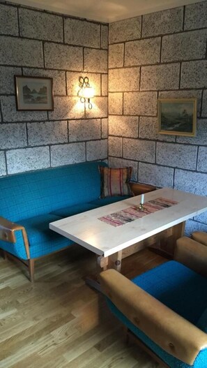 A cozy corner with a blue cushioned bench, a wooden table with a magazine, and a stone wall with framed pictures.