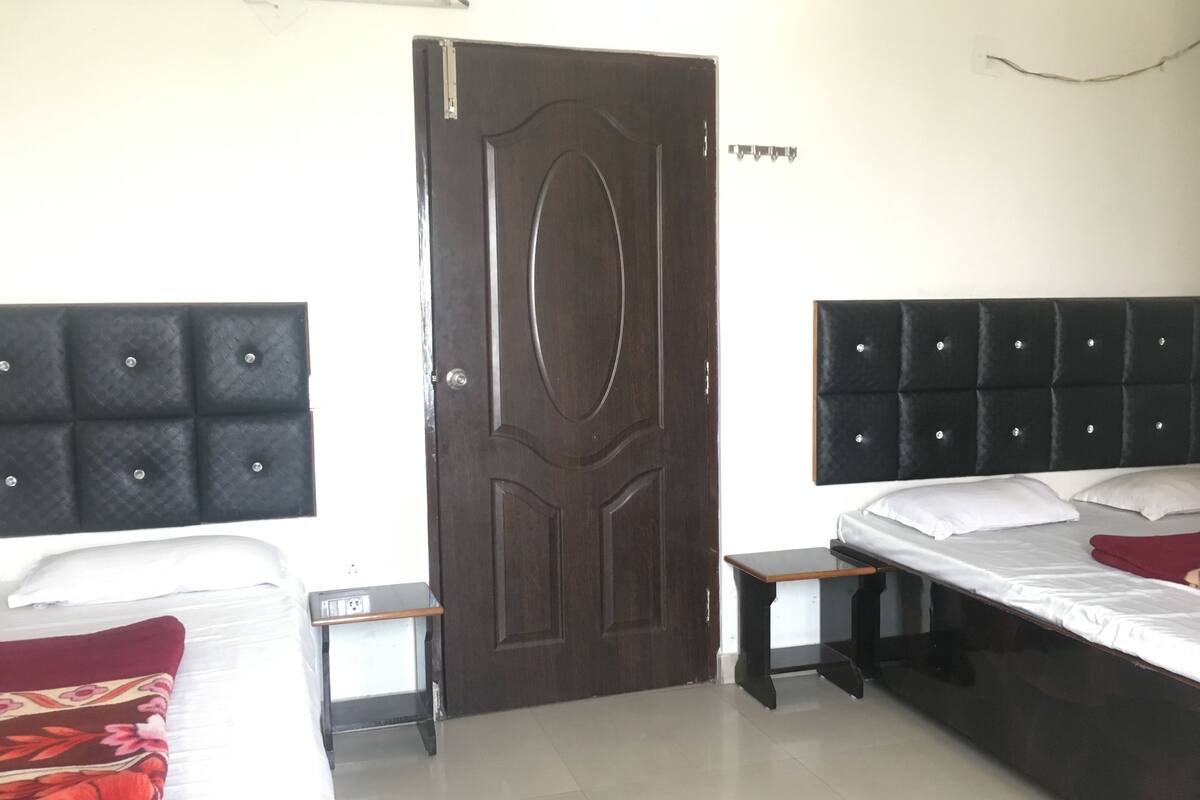 Quarto com duas camas, uma porta de madeira escura e uma pequena mesa de cabeceira com abajur.