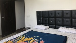 A bedroom with a dark headboard, a bed with floral bedding, a bedside table, and a door. - Hotel Sunny International (Jammu)