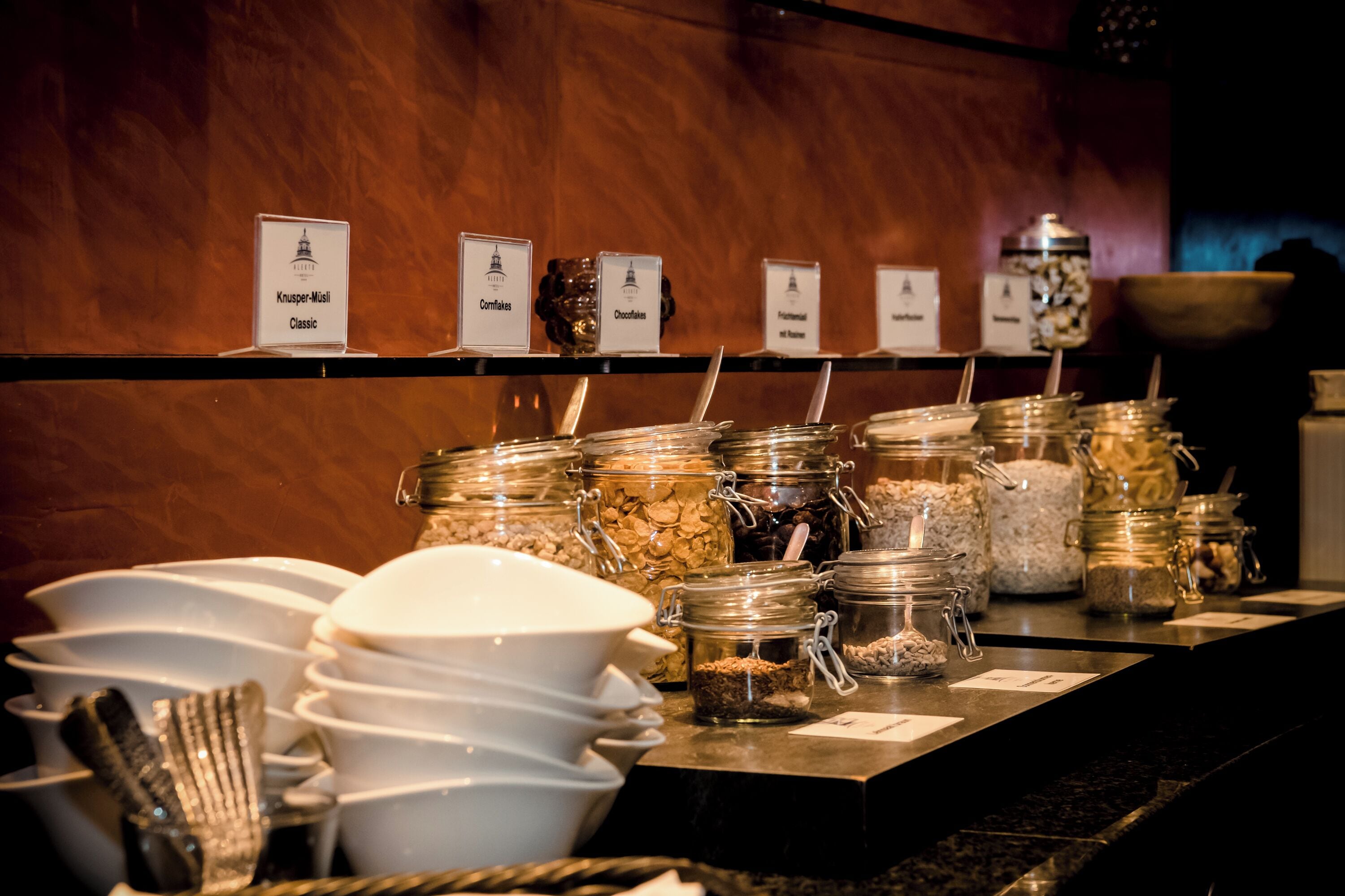 A buffet area with labeled food stations including Knusper-Mosaii Classic, Cornflakes, and Oatmeal.