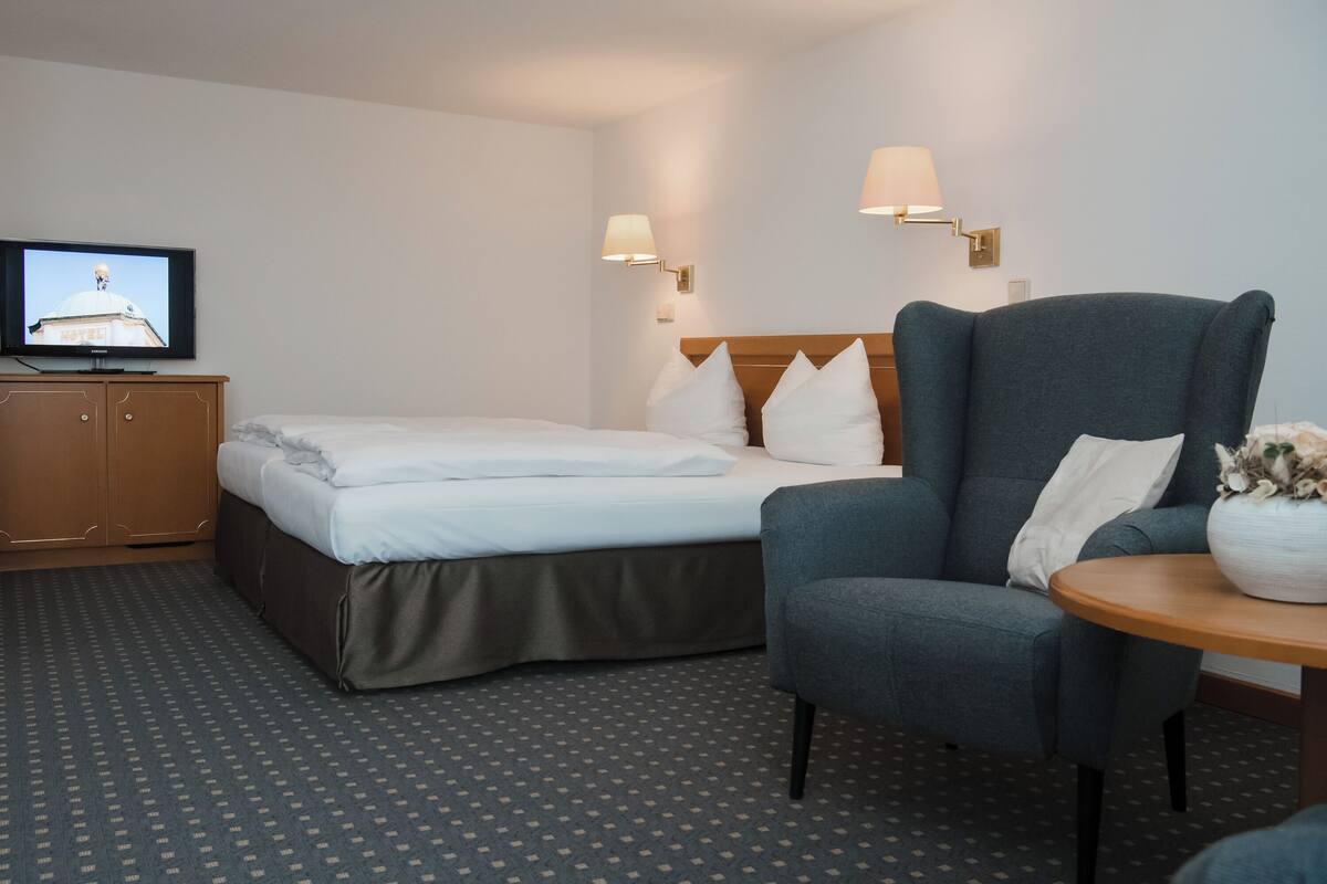 Quarto de hotel com cama, poltrona, mesinha, televisão e luminárias na parede.