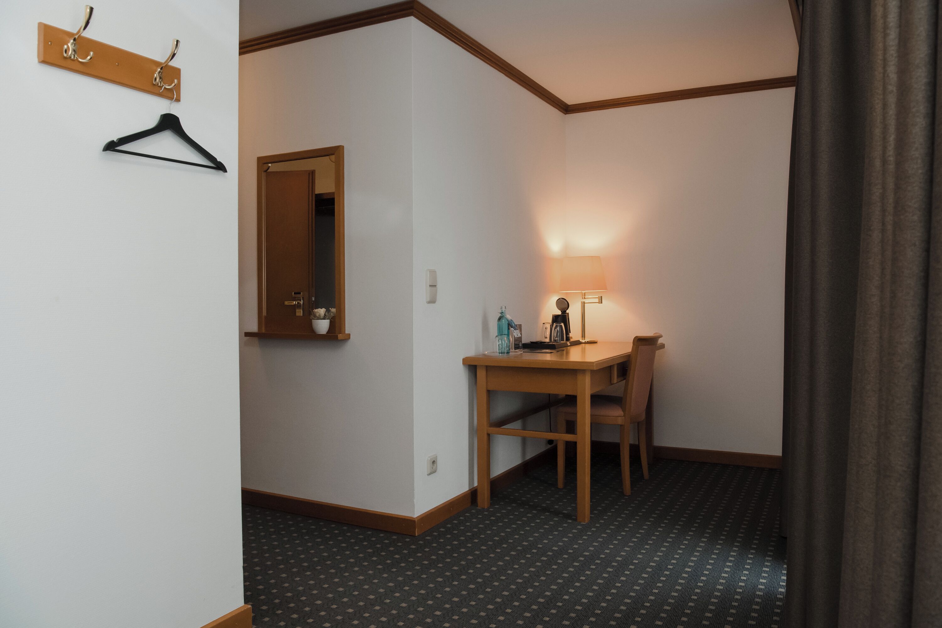 A hotel room with a wooden desk, a chair, a lamp, a coat rack, and a mirror.