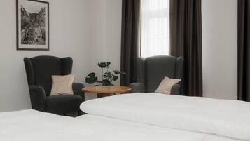 Habitación de hotel con dos sillones oscuros, una mesa redonda de madera y una cama con ropa blanca.