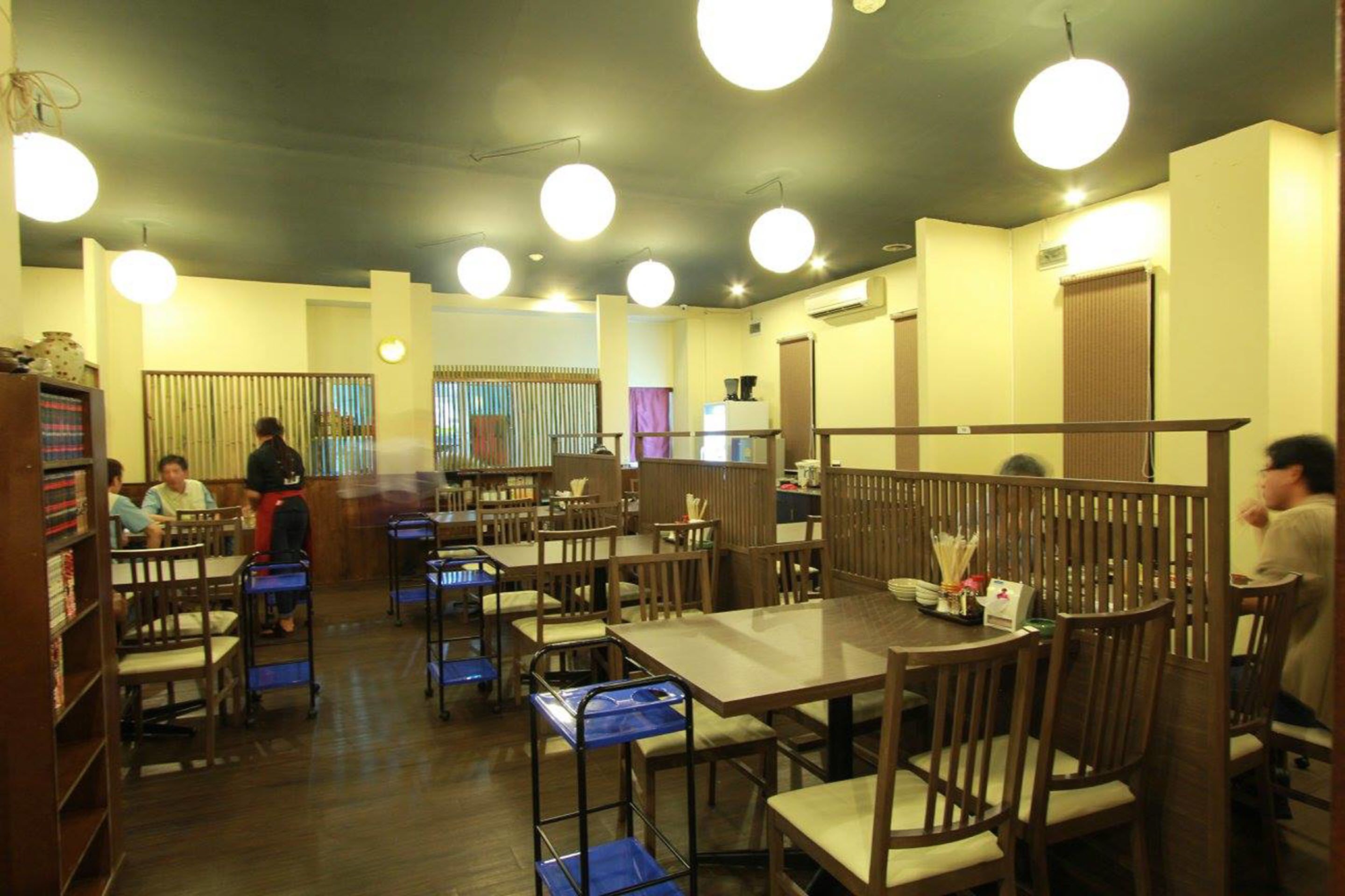 A restaurant with wooden tables and chairs, some with blue trays. There are round ceiling lights.