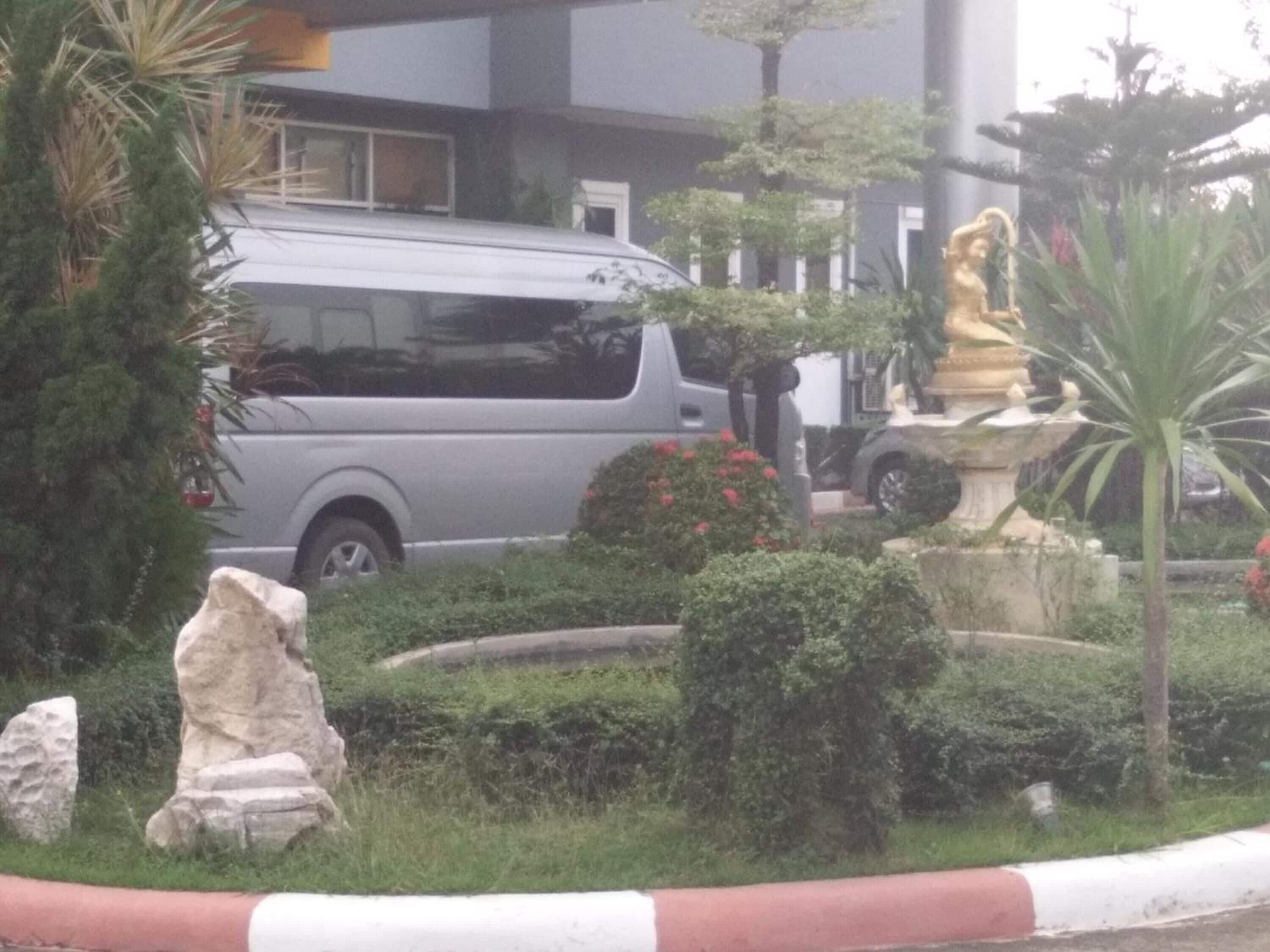 Un estacionamiento con una camioneta plateada, una estatua y un jardín cuidado con rocas y arbustos.