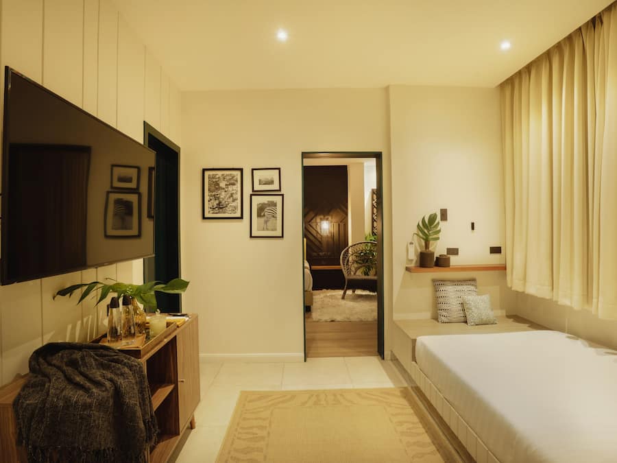 Une chambre moderne équipée d’un téléviseur à écran plat, d’un lit, d’une table de chevet agrémentée d’une plante et d’une porte donnant accès à une salle de bain.