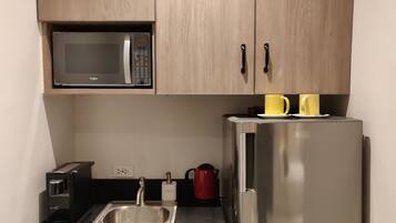 Una cocina compacta con microondas, un refrigerador de acero inoxidable, un lavamanos, una cafetera y dos tazas amarillas sobre el refrigerador.