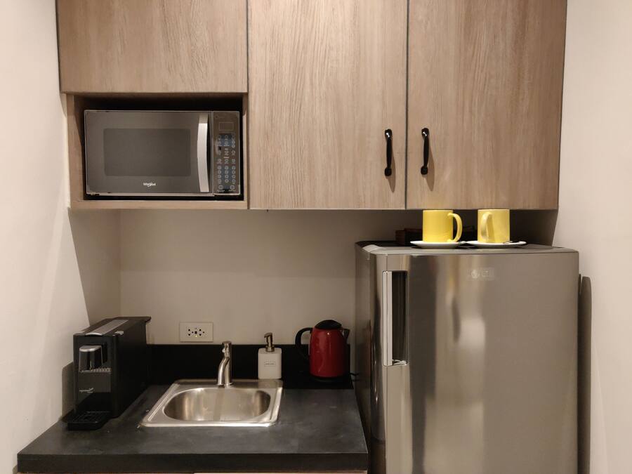 Una cocina compacta con microondas, un refrigerador de acero inoxidable, un lavamanos, una cafetera y dos tazas amarillas sobre el refrigerador.