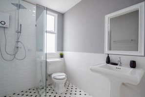 A bathroom with a glass-enclosed shower, a white toilet, a sink with a mirror above it, and a window providing natural light. - Wan Arun Houses (Hua Hin)