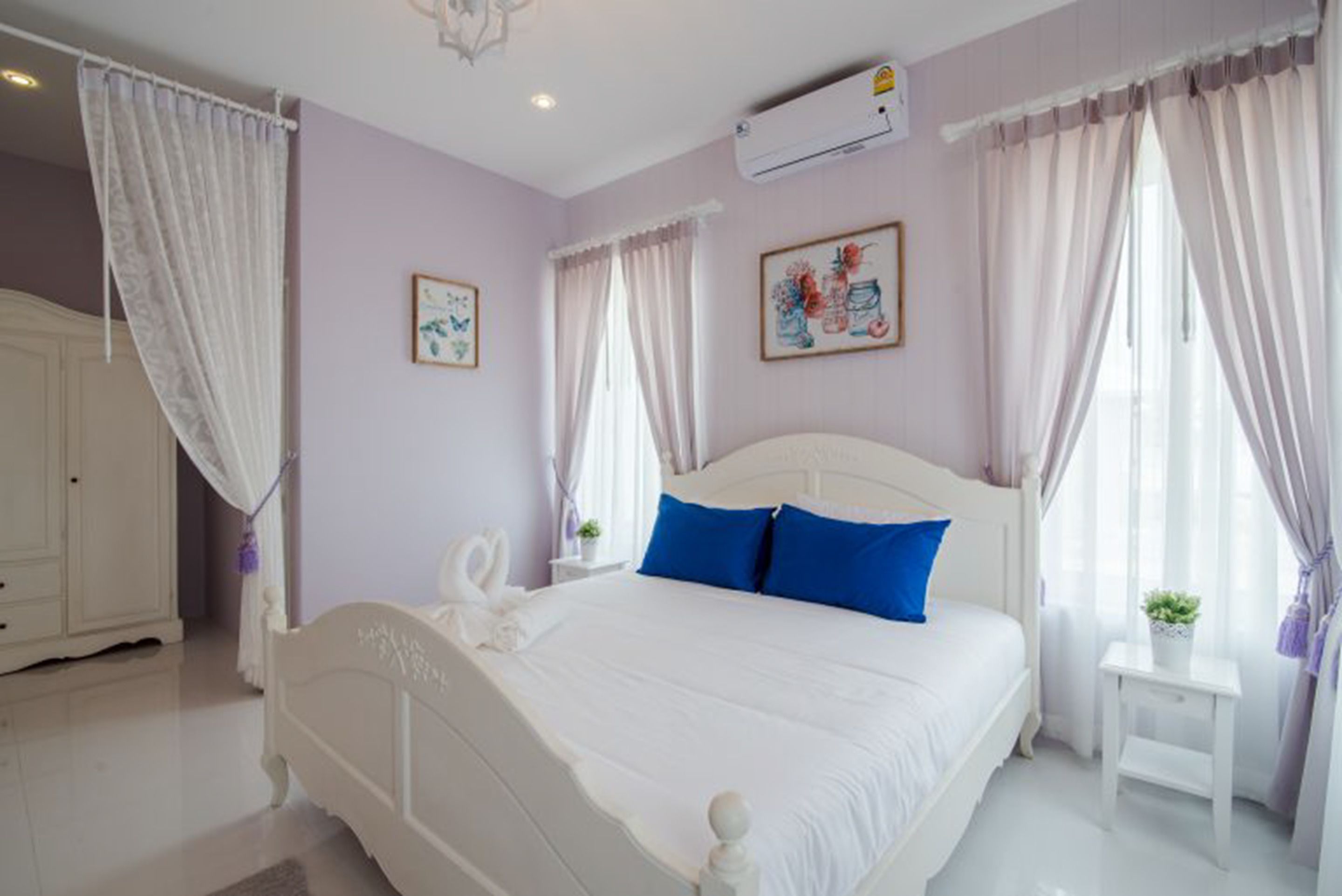Un dormitorio con cama blanca, almohadas azules, armario blanco y dos aires acondicionados instalados en la pared.