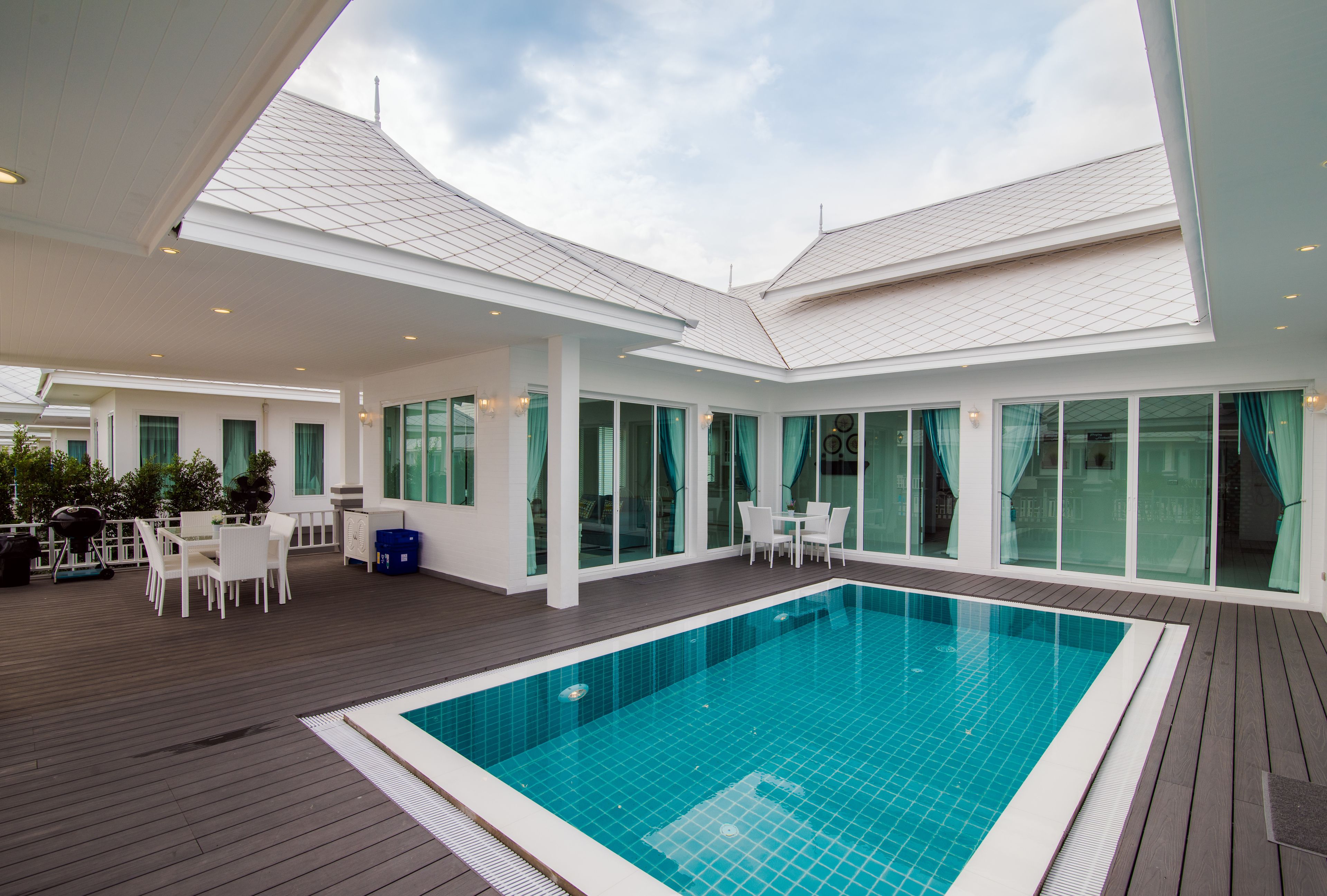 Un área de piscina exterior moderna con un deck de madera, mobiliario blanco y ventanas amplias.
