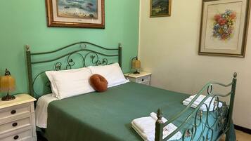 A neatly made bed with a green bedspread, white pillows, and a brown accent pillow. There are two framed artworks on the walls, a lamp on the nightstand, and a small drawer unit beside the bed.