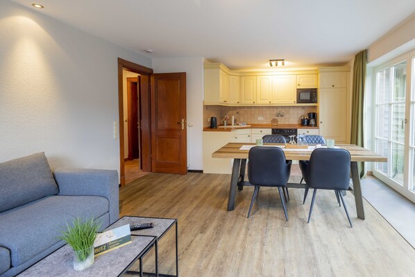 A modern living room with a grey sofa, a coffee table with a remote control, and a small plant. A dining area with a wooden table and four chairs is visible. The kitchen area is equipped with light-colored cabinets and a microwave. - Butz (Scharbeutz)