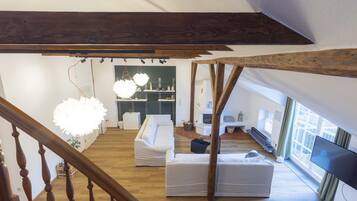 A spacious living area with a wooden staircase, white sofas, a large window, and a TV.