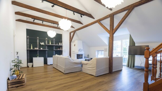 Un loft spacieux avec de hauts plafonds, des poutres apparentes et un grand salon doté de deux canapés blancs et d’un plancher en bois.