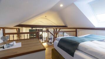 Un dormitorio con techo inclinado, vigas de madera, una cama con ropa de cama blanca y aguamarina, un escritorio de madera y una mesita con un florero y papeles.