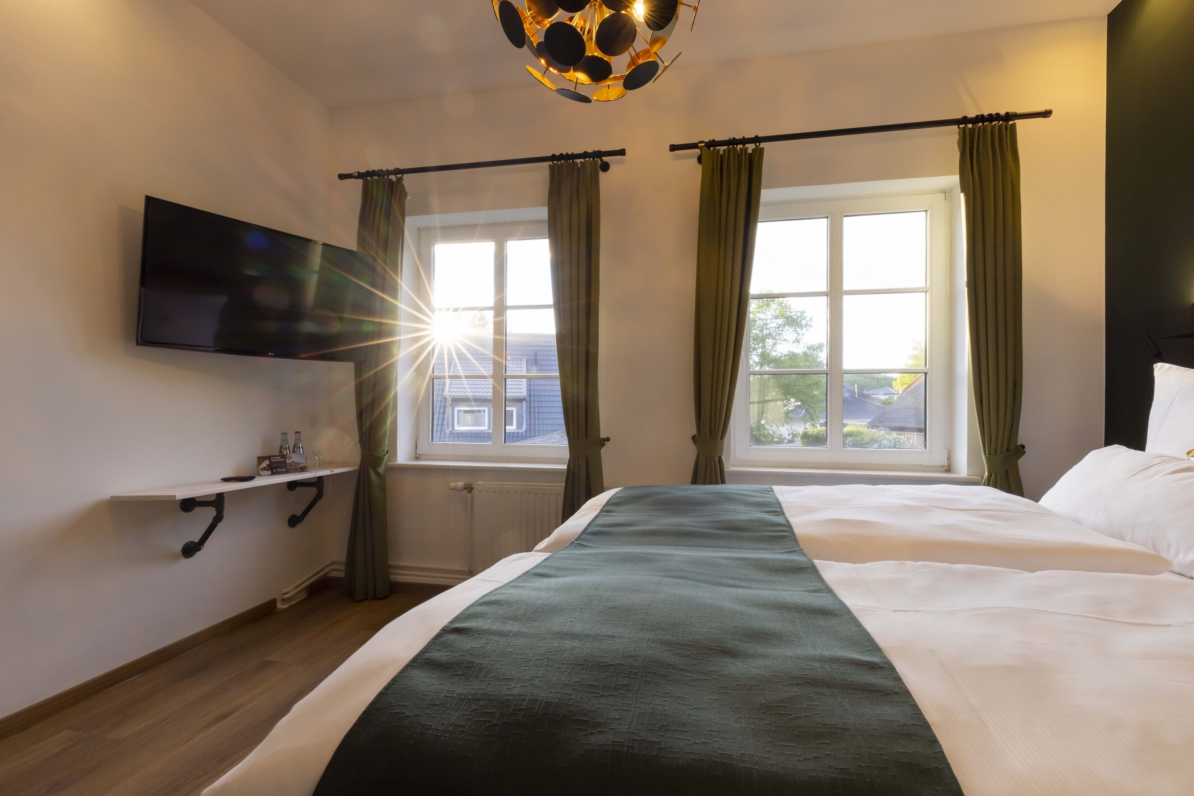 A hotel room with a bed, a TV mounted on the wall, a window with curtains, and a shelf with bottles.