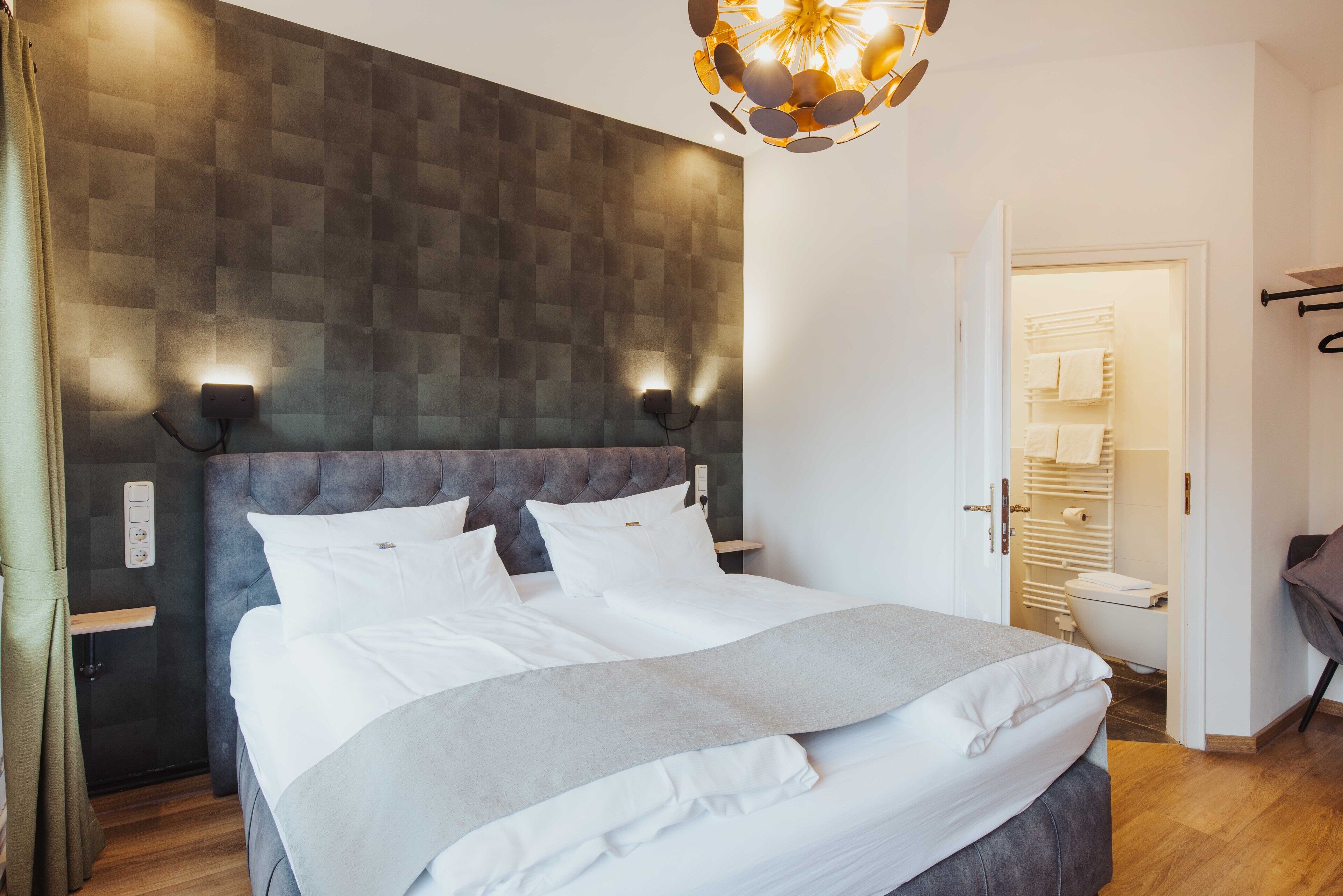 Une chambre à coucher moderne avec une tête de lit capitonnée, un grand lit avec du linge de lit blanc, un plaid gris et un luminaire fixé au mur.