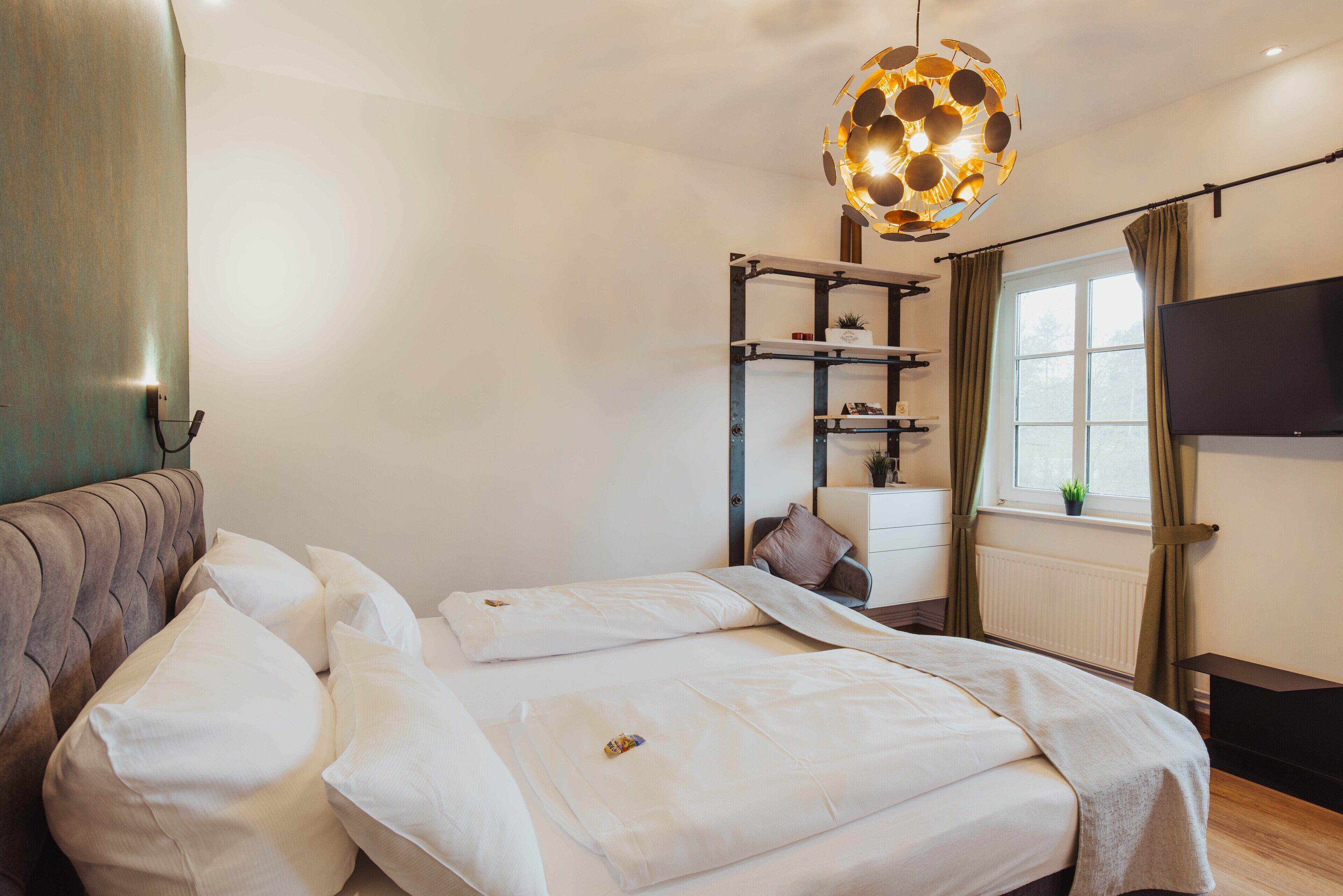 Un dormitorio con cama, mesita de noche, televisor, ventana con cortinas y lámpara colgante.