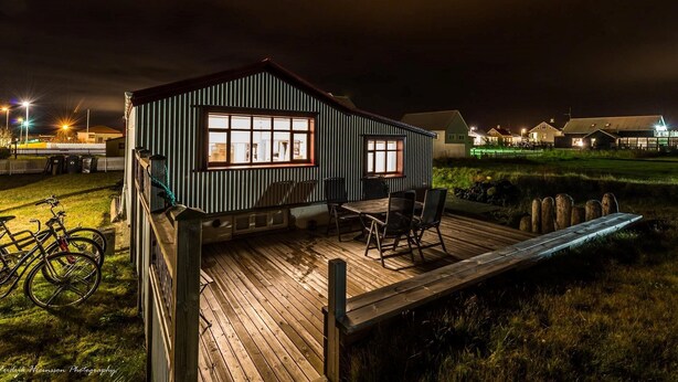 A wooden deck with a table and chairs, a bicycle, and a building with windows. - Sea Side Cottages (Eyrarbakki)