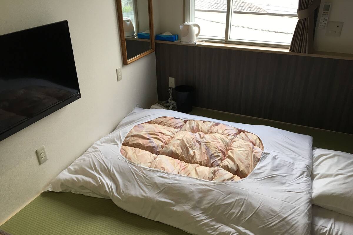 A bed with a patterned duvet, a mounted television, a window with curtains, and a bedside table with a kettle.