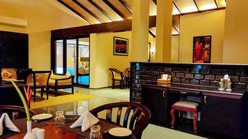 A dining area with a wooden table set for two, a bar area with a brick backsplash, and a view of a room with a bed and curtains.