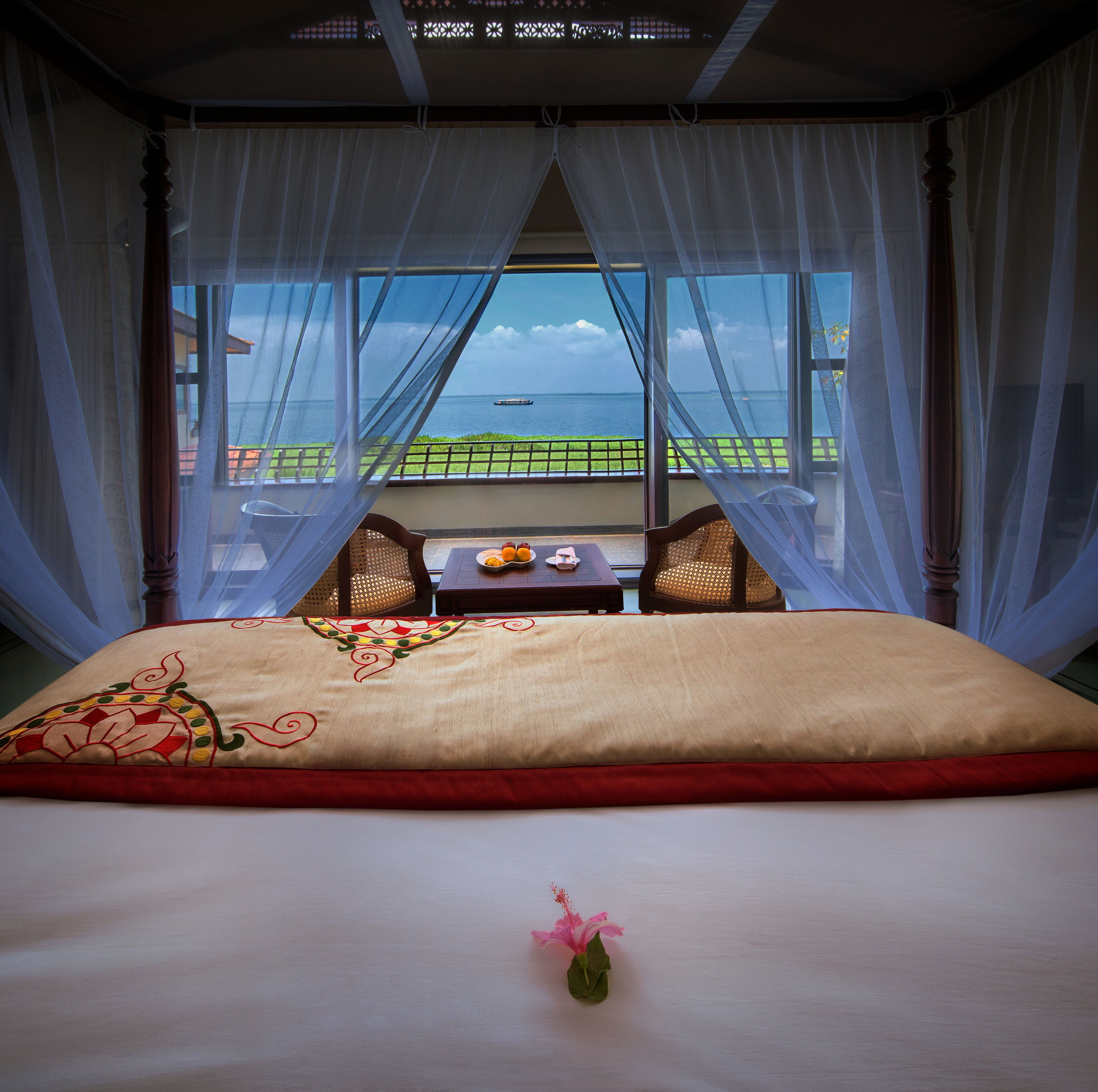 Una cama con vista al mar, una mesa con fruta y un jarrón con una flor.
