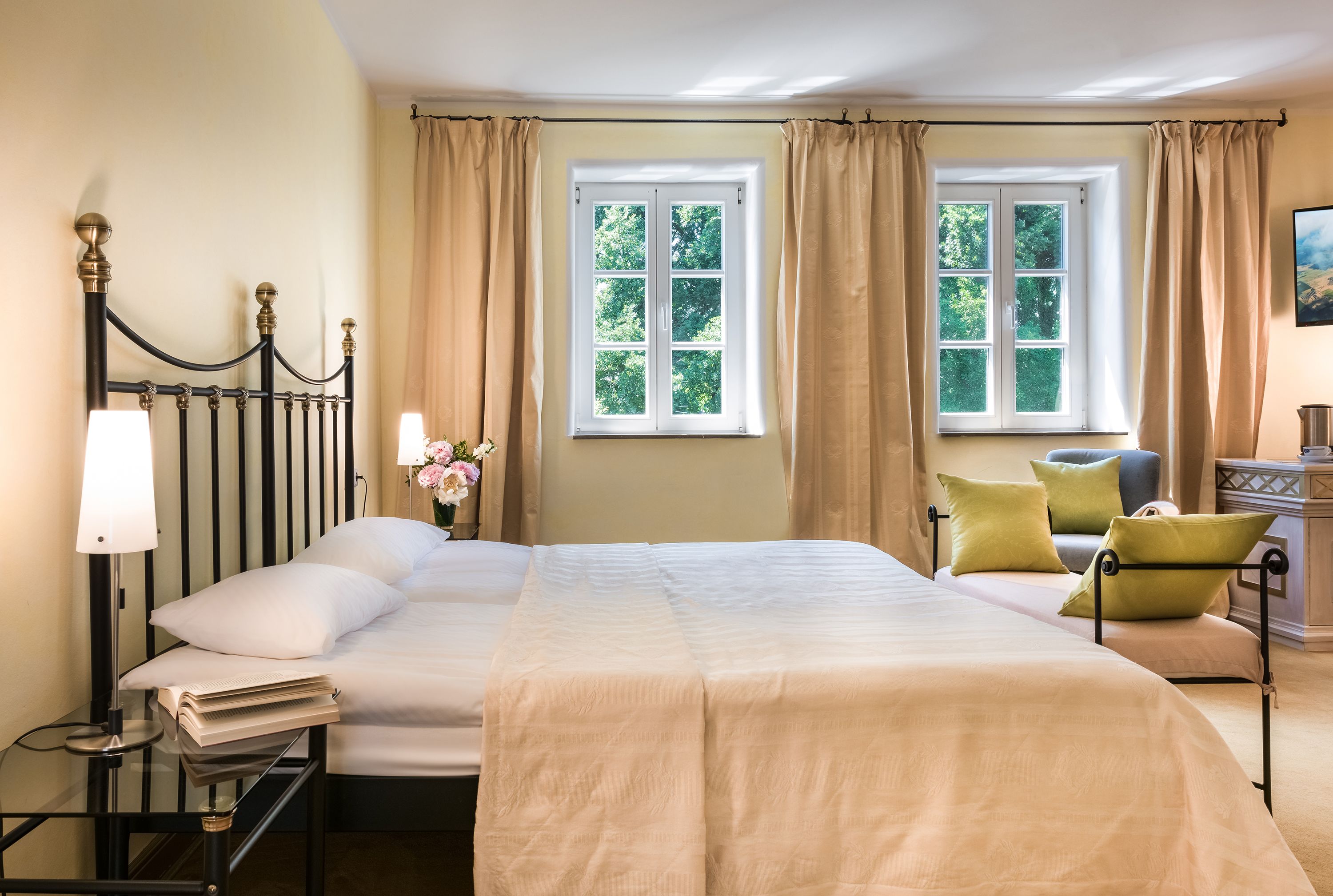 A hotel room with a large bed, a bedside table with a lamp, a chair, and two windows with curtains.
