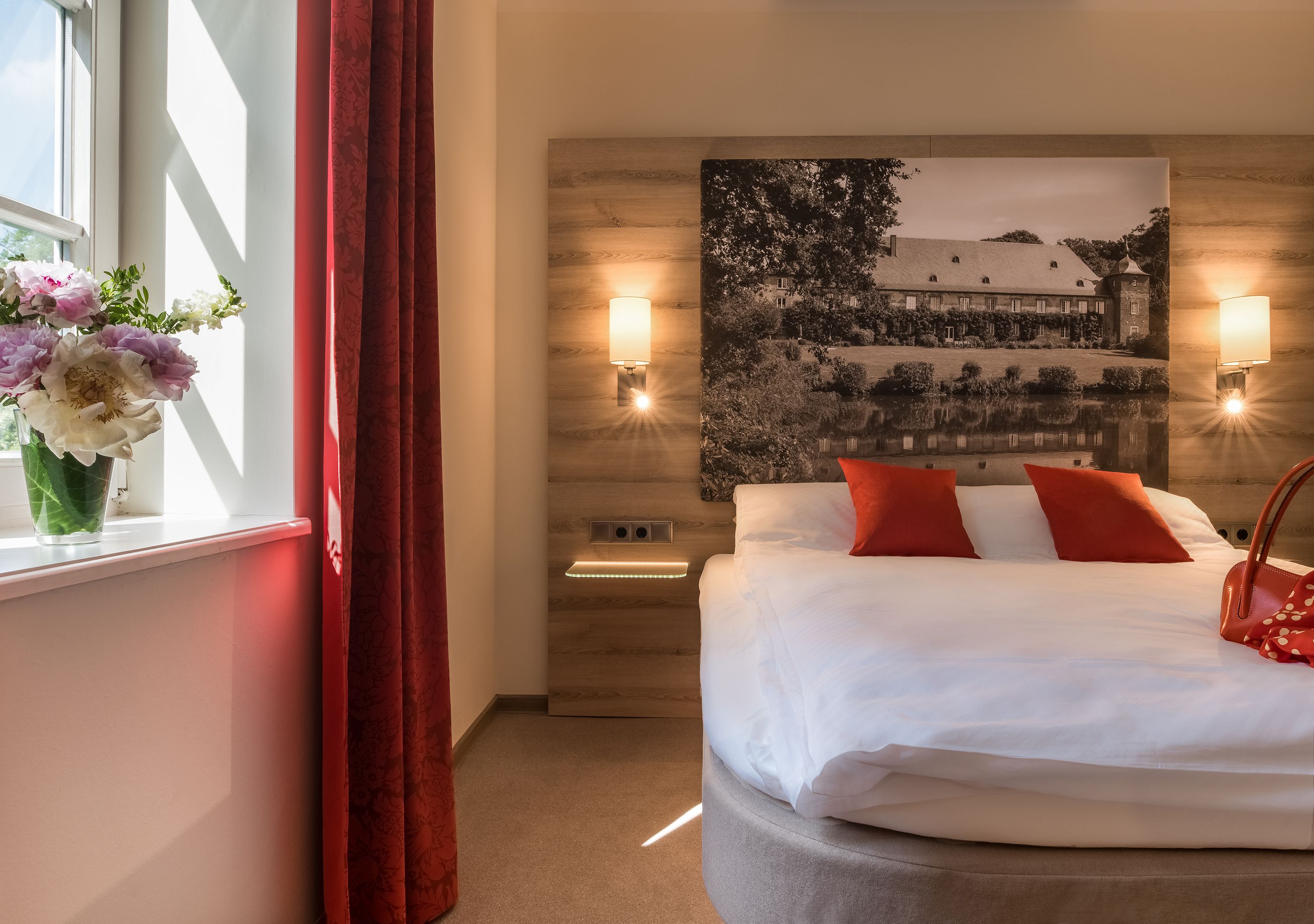 Habitació d'hotel amb un llit, una tauleta de nit, una làmpada, un vas amb flors i una finestra amb cortines.