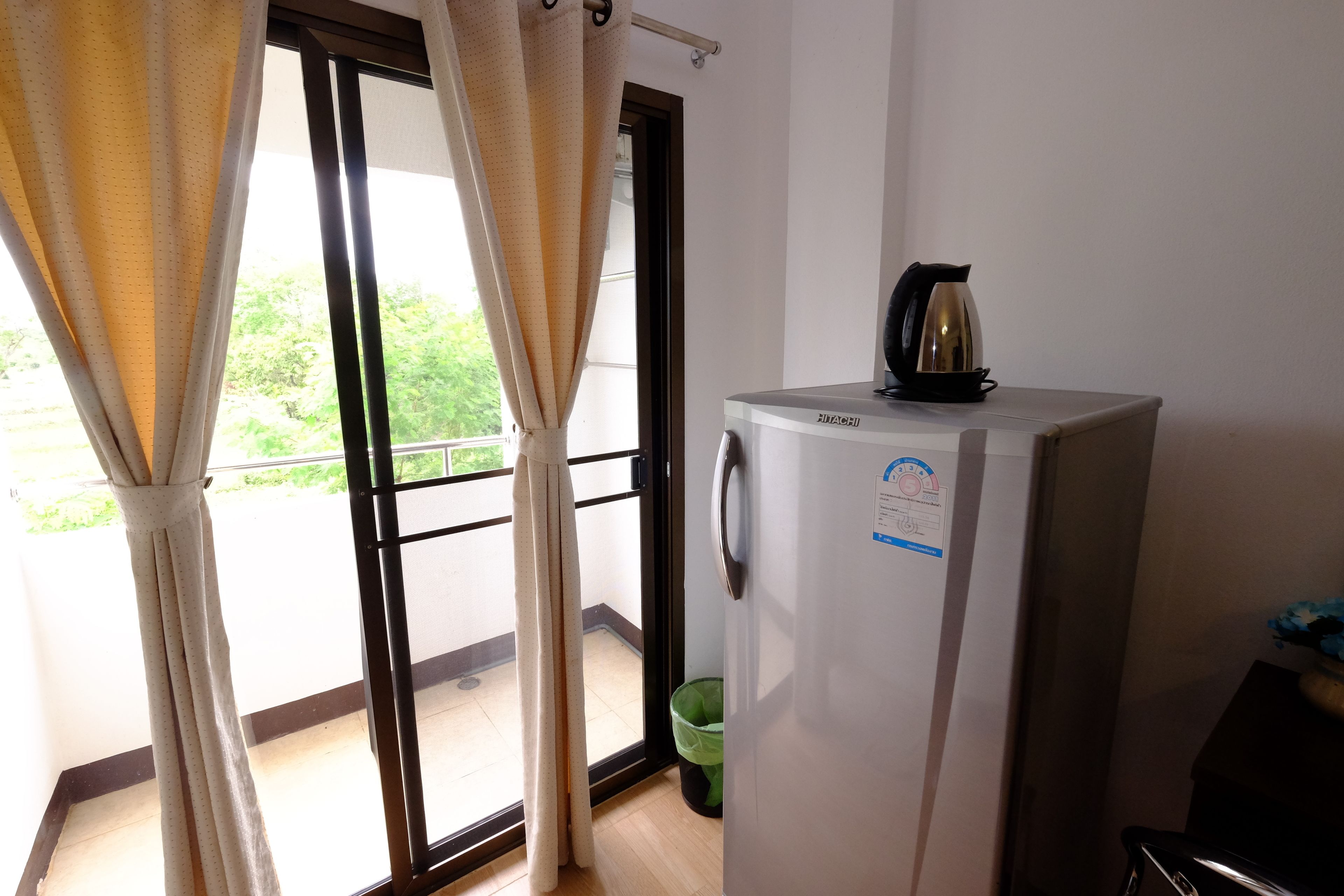 A room with a sliding glass door leading to a balcony, a refrigerator with a kettle on top, and a green trash bin.