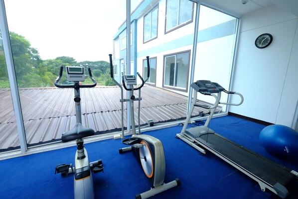 A gym with cardio equipment, a blue carpet, and a view of a wooden deck through large windows. - Tafah Residence (Sakon Nakhon)