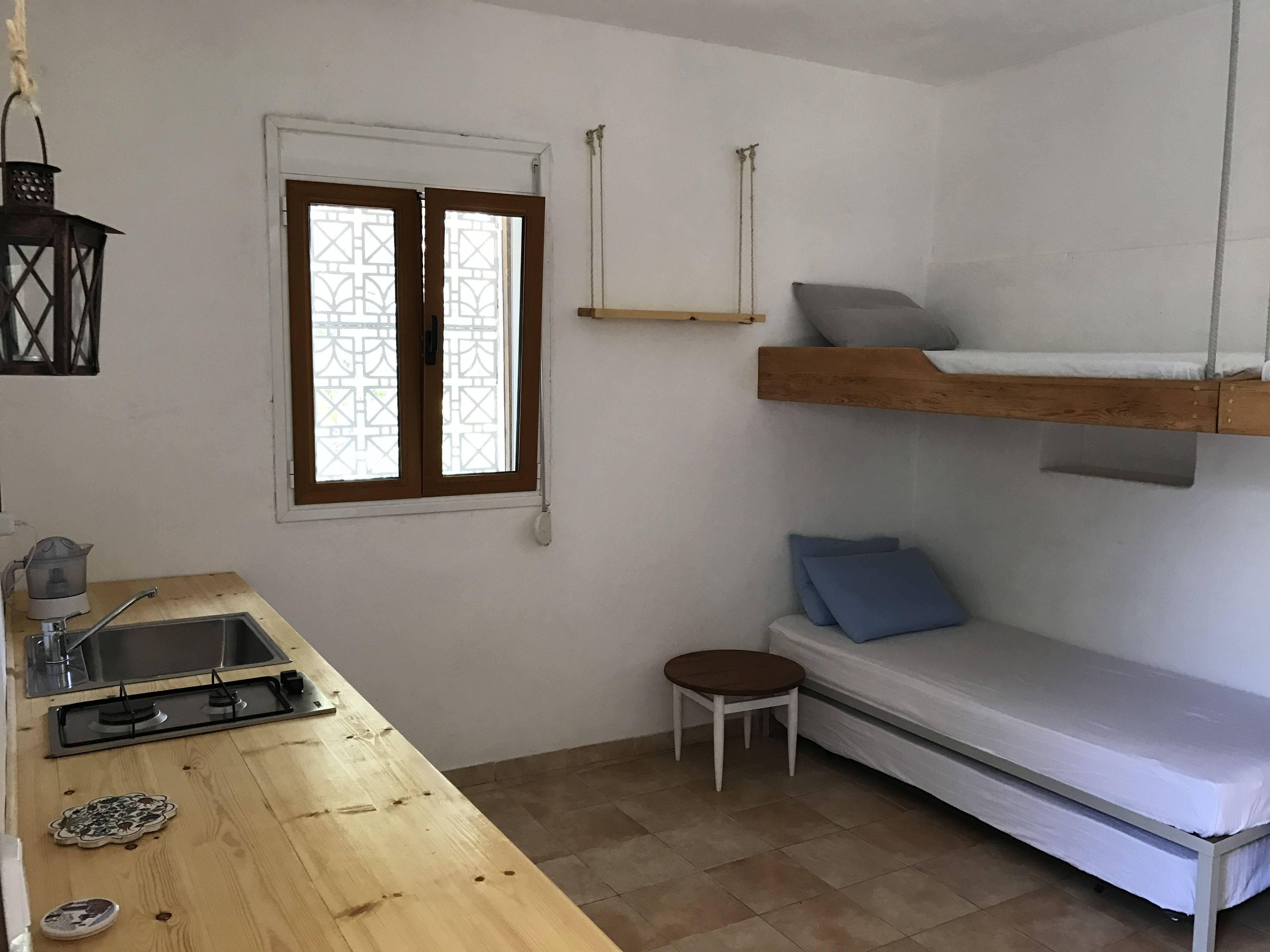 Ein kleines Zimmer mit Etagenbett, einem Tischchen, einem Waschbecken und einem Fenster mit schmiedeeisernem Gitter.