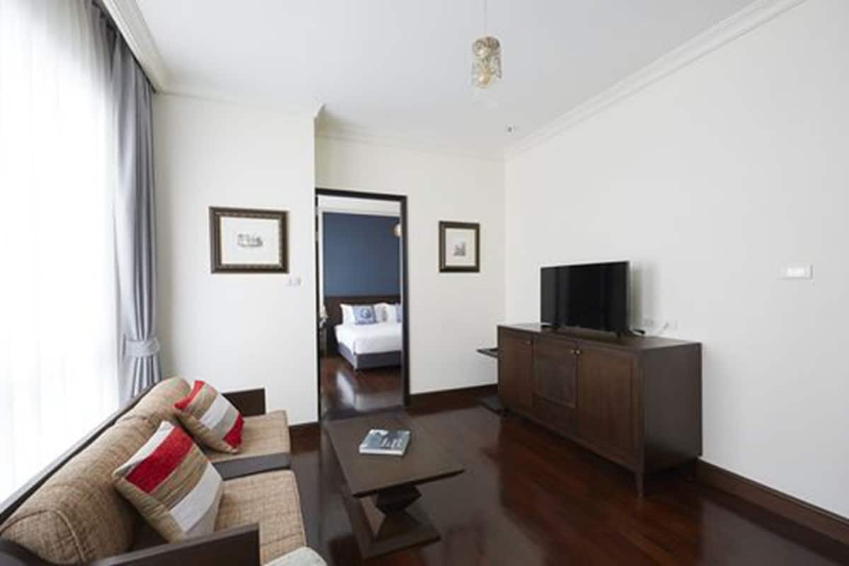A modern hotel room with a sofa, a coffee table, a TV, and a wardrobe. There is a door leading to a bedroom with a bed and a window with curtains.