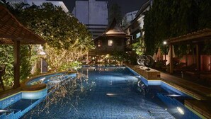 Une piscine nocturne avec une fontaine, entourée d’une végétation luxuriante et bordée d’édifices à l’architecture traditionnelle.