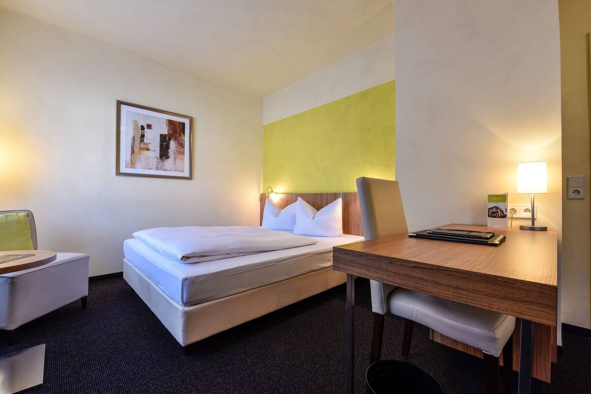 A hotel room with a bed, a desk, a chair, a lamp, and a framed picture on the wall.