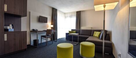 A modern hotel room with a kitchenette, a sofa, two yellow ottomans, a desk, and a TV.
