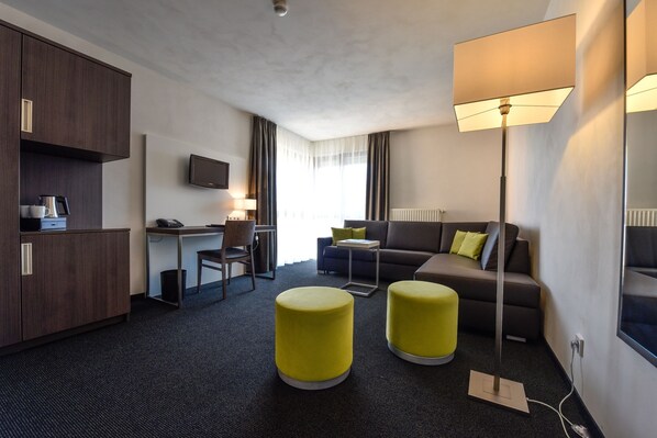 A modern hotel room with a kitchenette, a sofa, two yellow ottomans, a desk, and a TV.