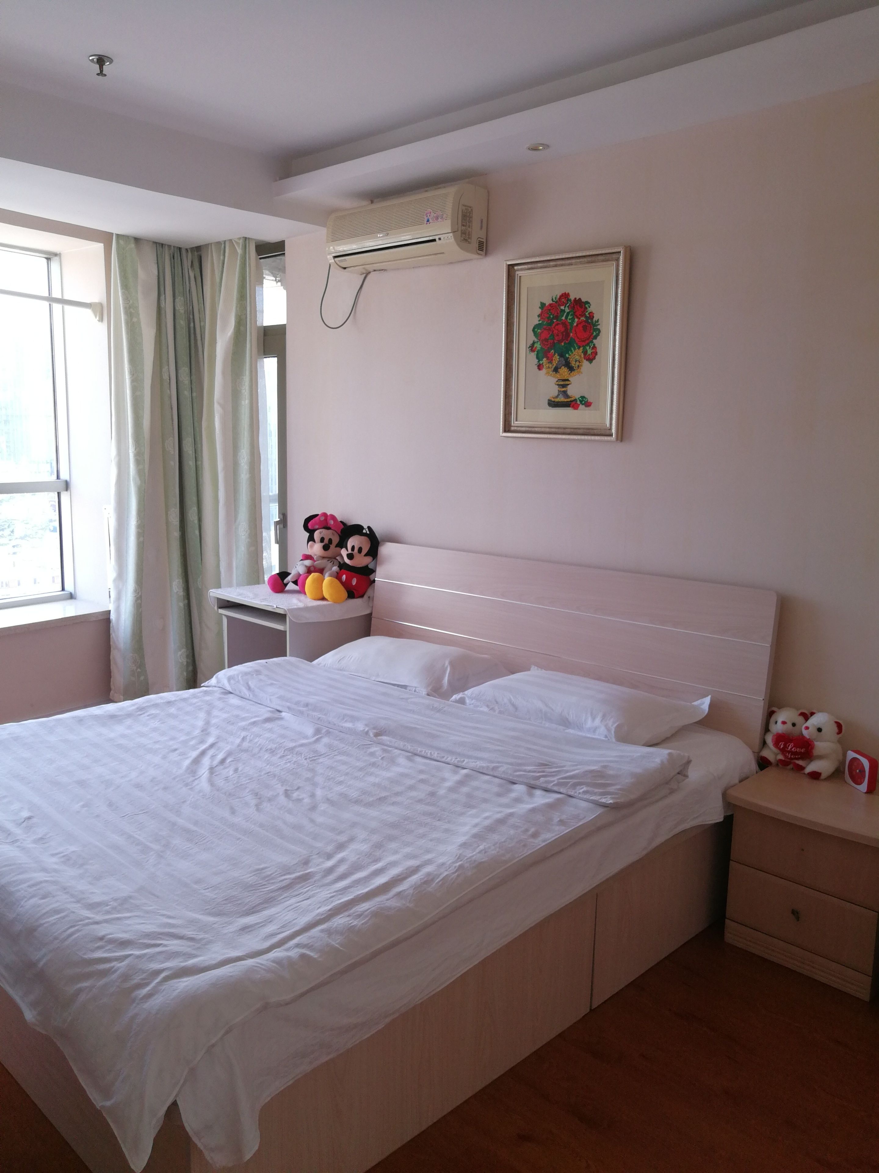 Een slaapkamer met een bed, een nachtkastje, een wandairconditioner, een raam met gordijnen en een ingelijste foto aan de muur.